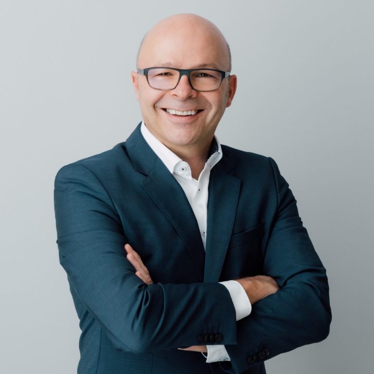 Porträt Simon Hauger Mann in Anzug mit Brille, lächelt und steht mit verschränkten Armen.