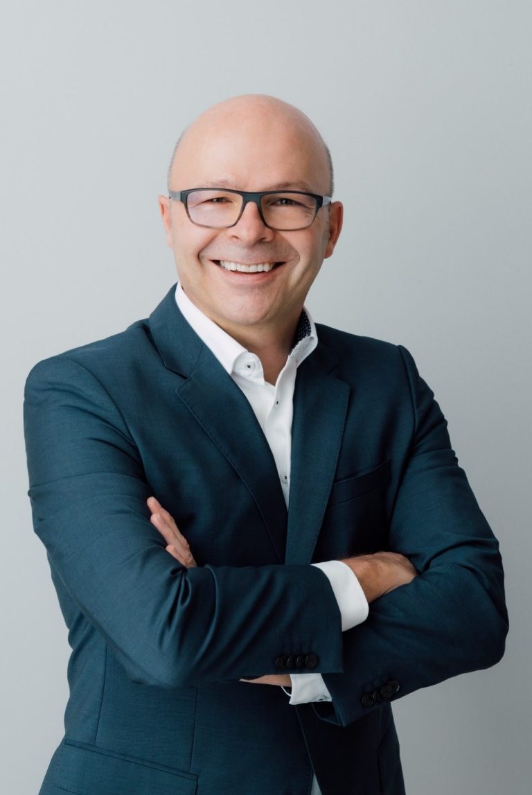 Mann mit Glatze und Brille in Anzug, lächelt und steht mit verschränkten Armen.