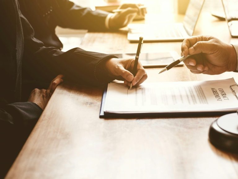 Hände beim Unterzeichnen eines Dokuments auf einem Tisch, mit Laptop im Hintergrund.